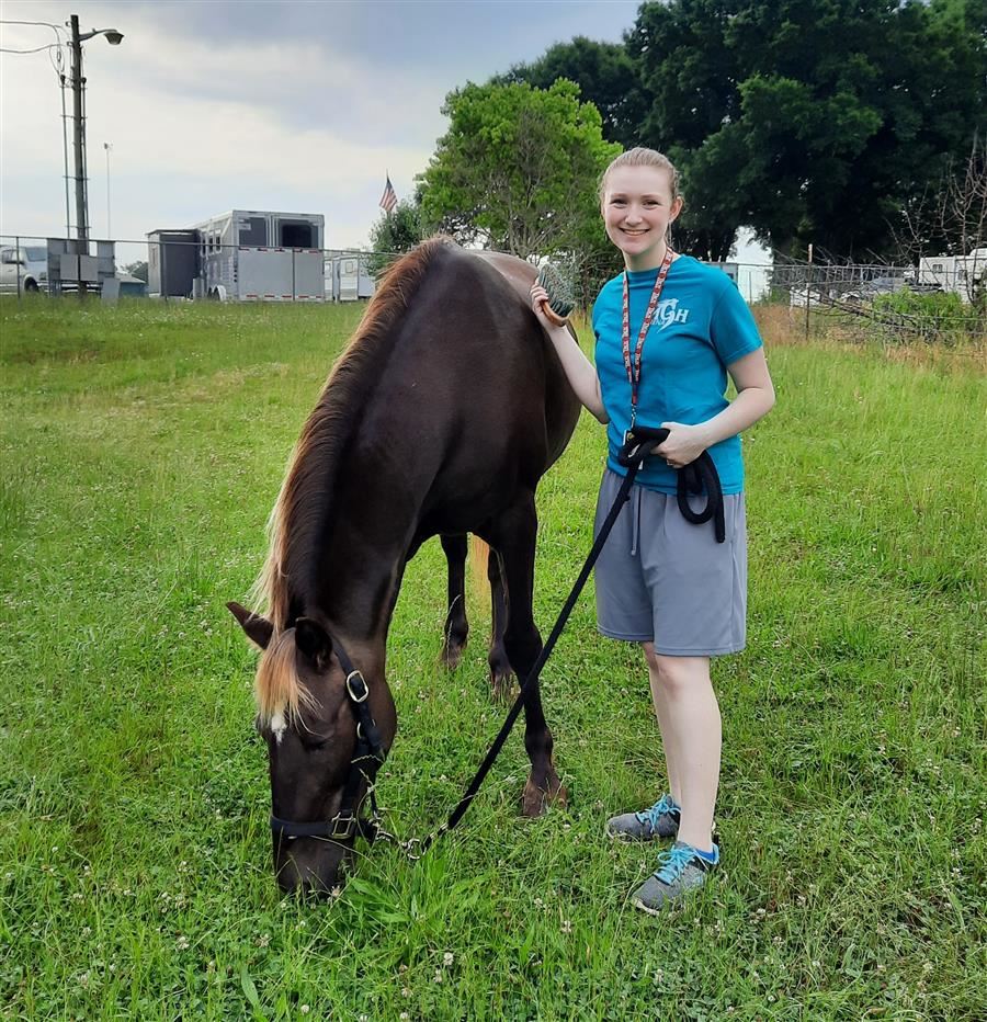 Alyson Linn smiles to camera. Next to her is a horse grazing in the grass. 